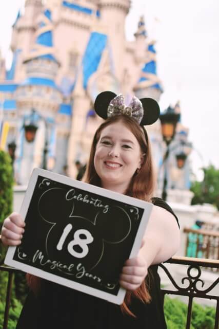 Girl in Mickey Mouse hat holding sign that says 18