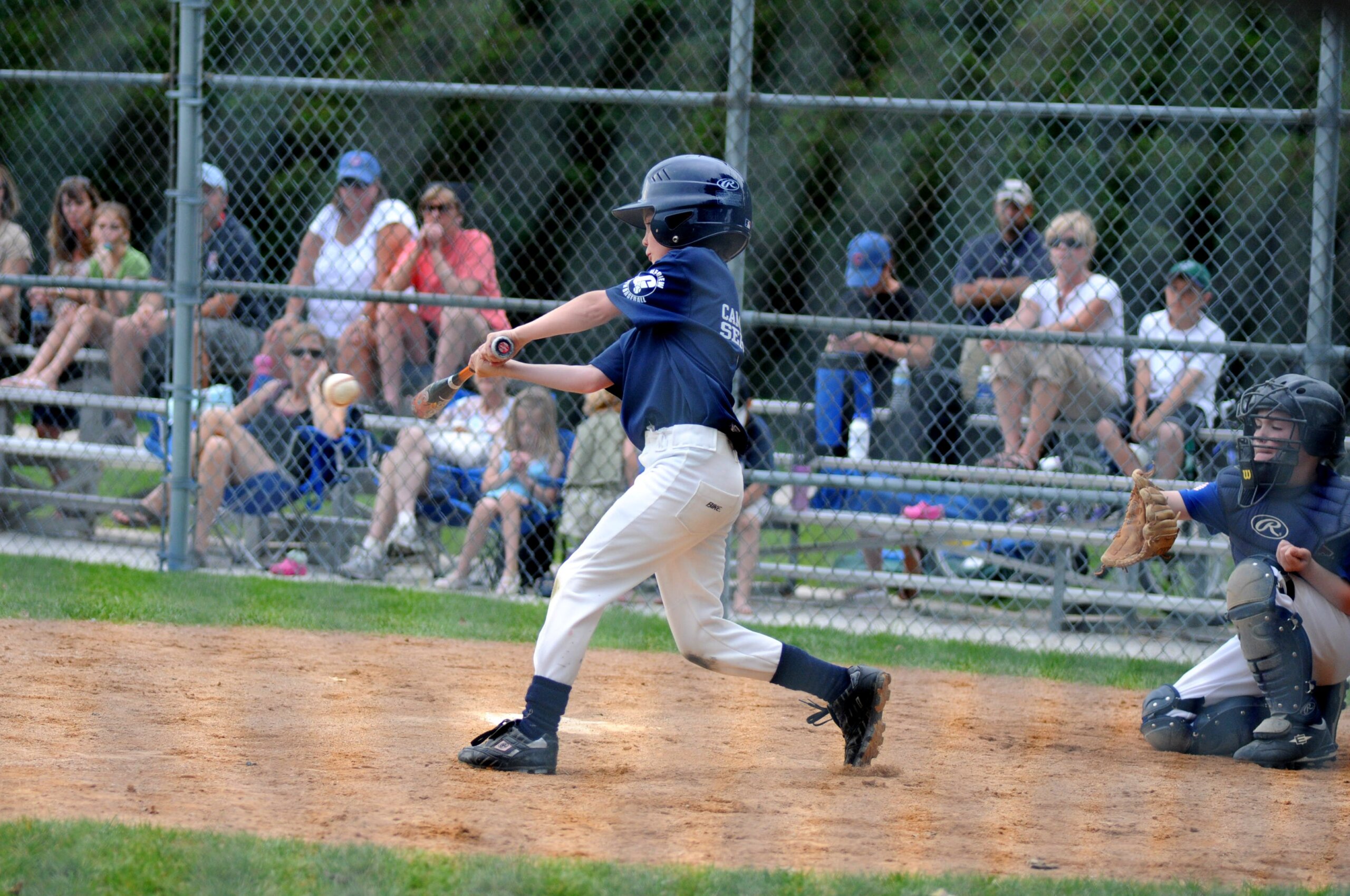 Young boy swinging a bat at a ball with onlookers in bleachers.