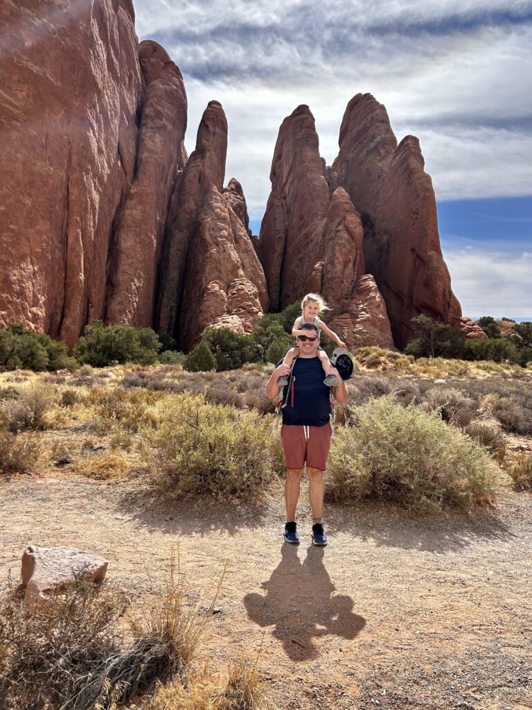 Man with girl on shoulders in front of red rocks.