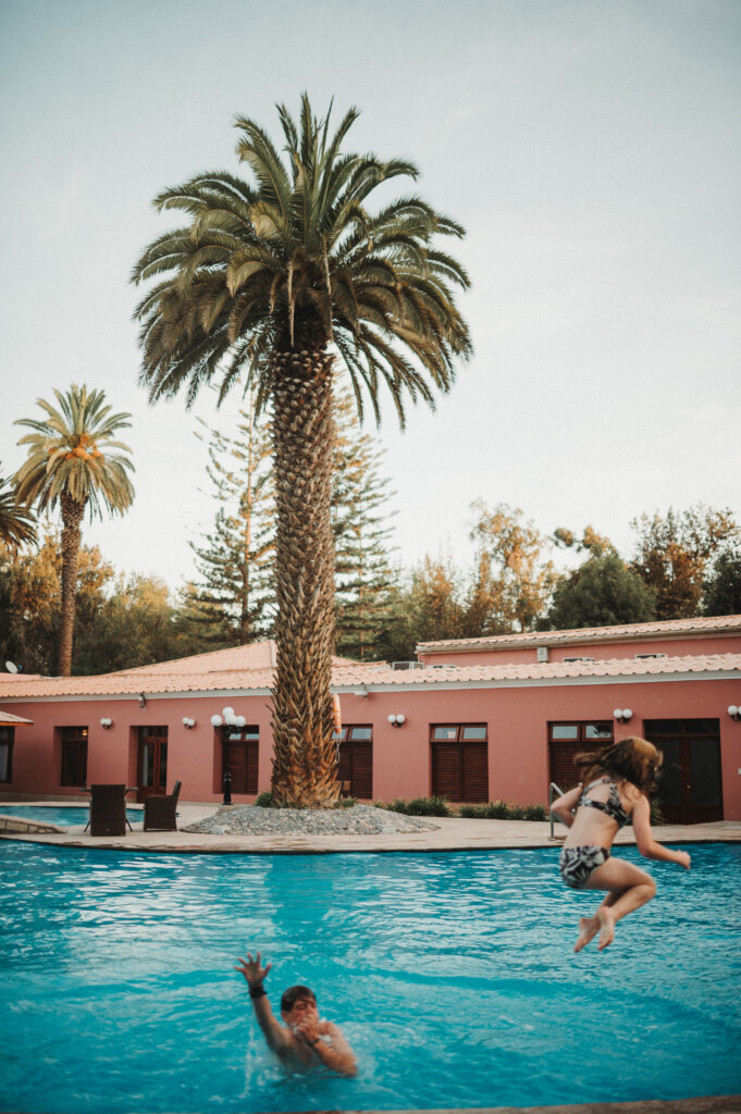 Children jumping into a large pool with a pink building and palm tree in the background.