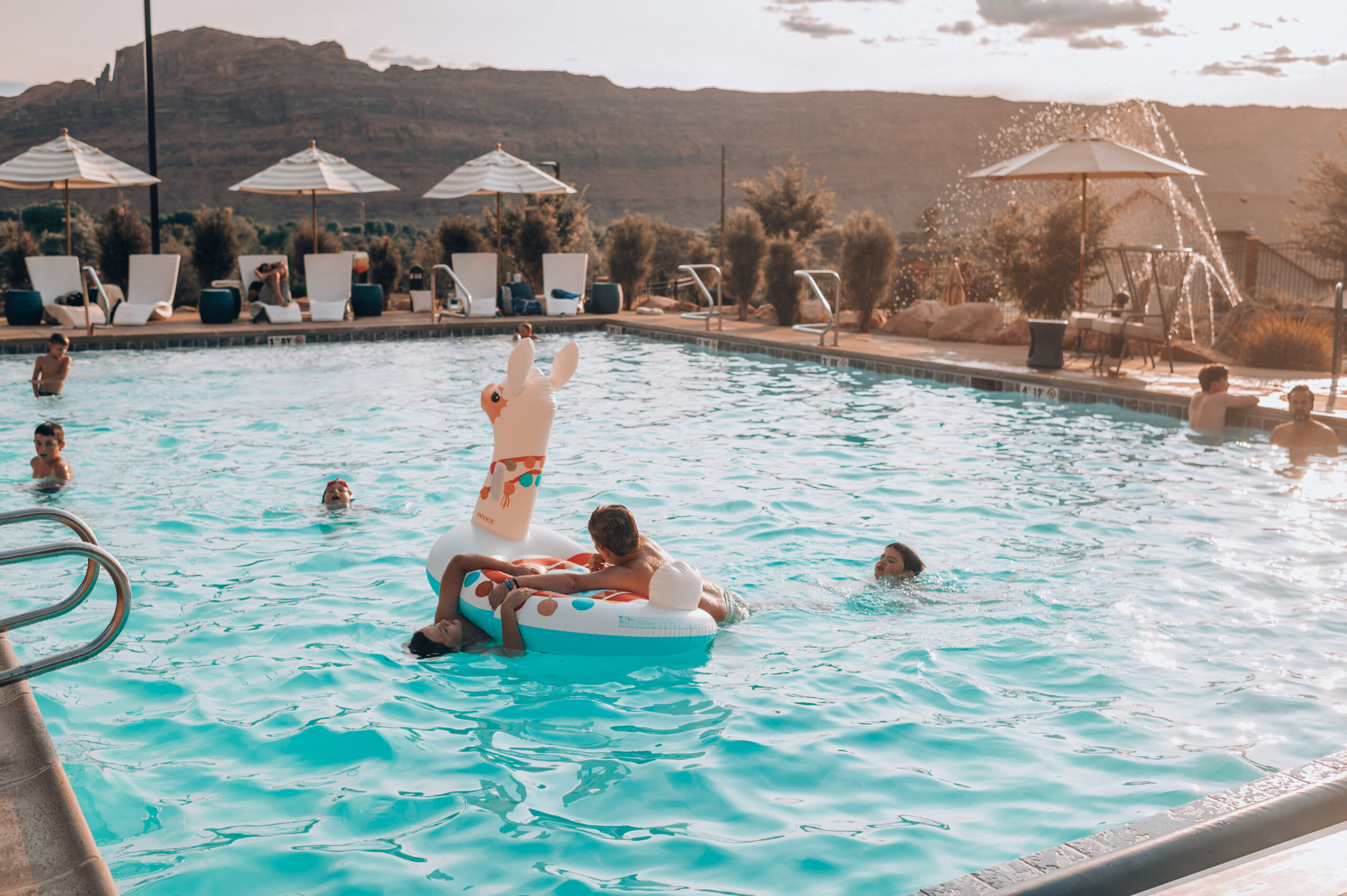 Blue pool with children swimming and playing on an inflatable llama with a desert background