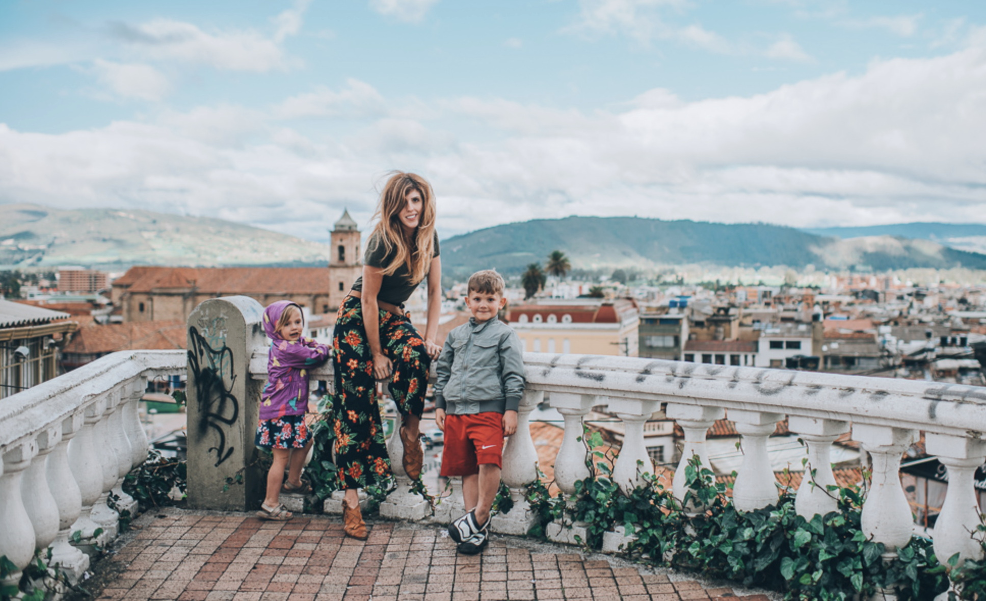A woman and two young children staning on a balcony with mountains in the background