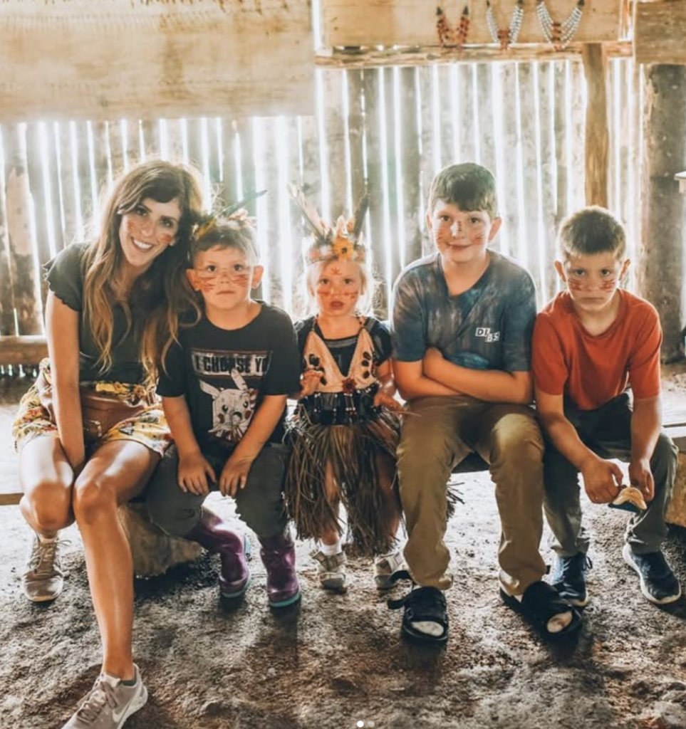 Four kids and a woman sitting on a bench inside a hut in the Amazon rainforest wearing traditional makeup and clothing.