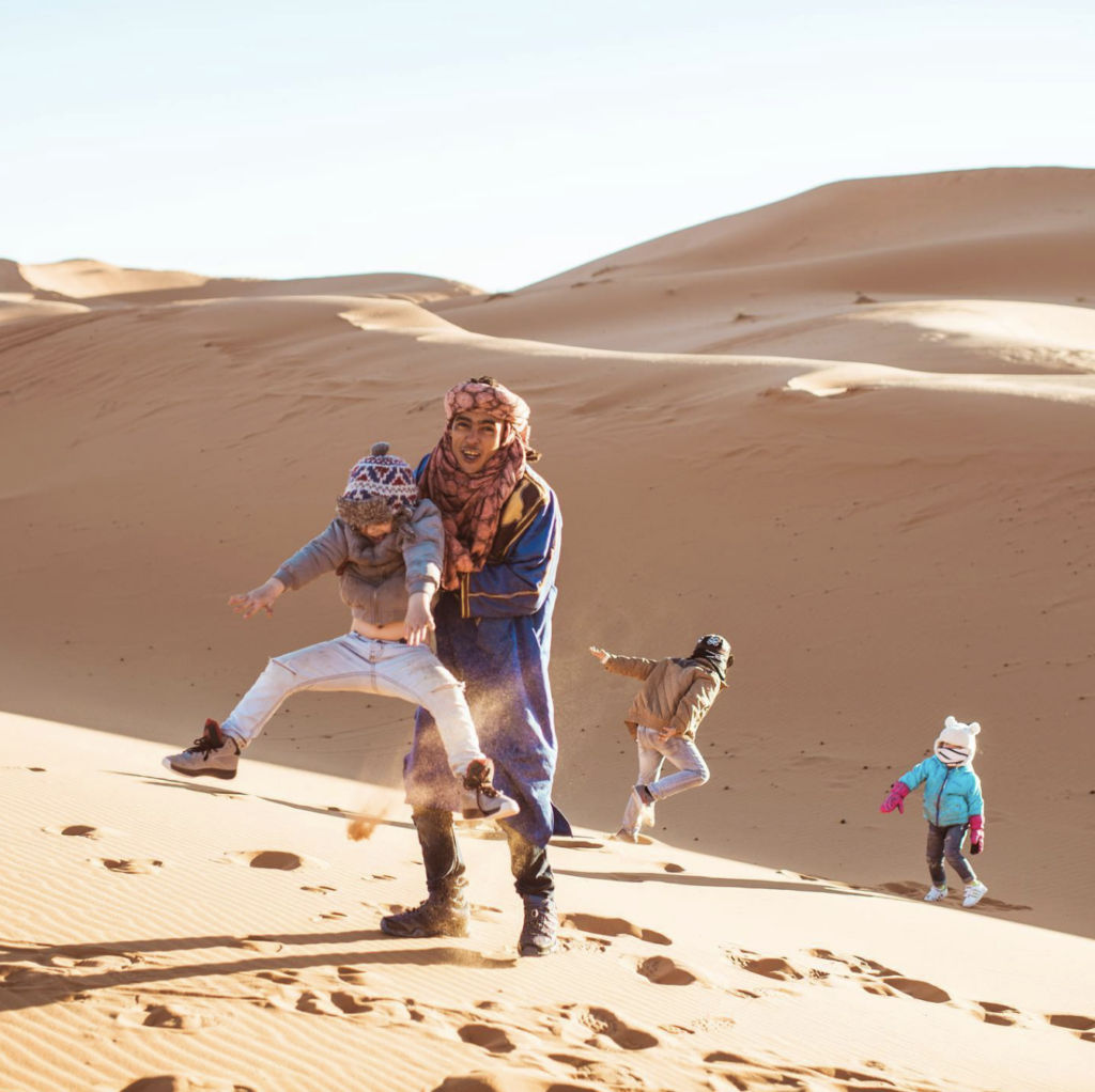 Three children and a nomad boy running around sand dunes in Morocco